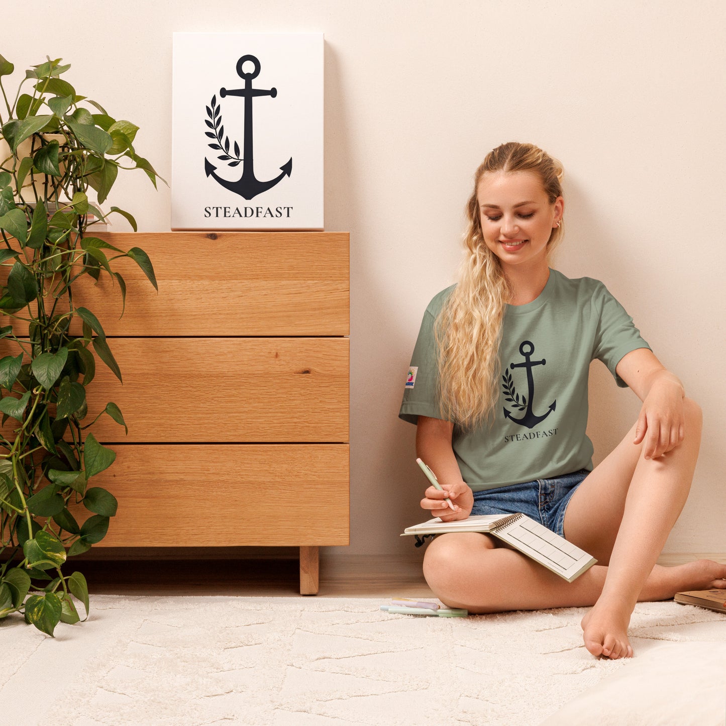 Young woman sitting on floor wearing Spirit Wear Christian anchor shirt for men and women in sage green Steadfast print